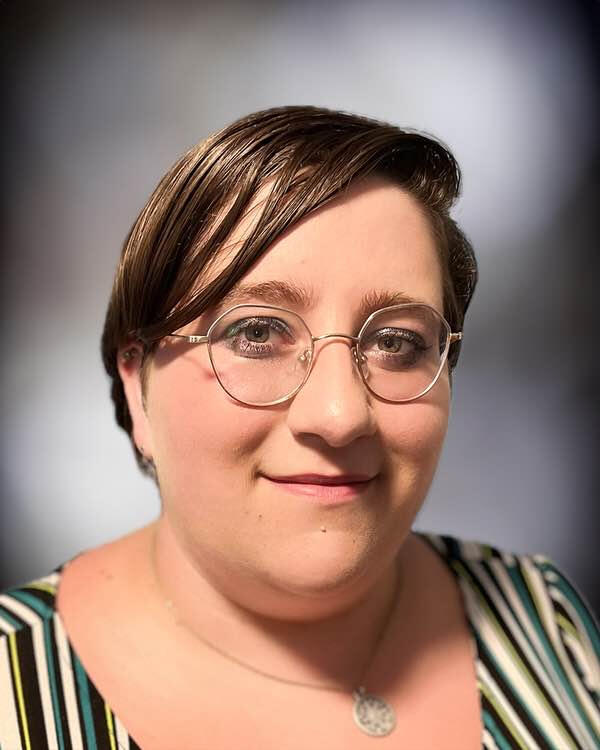 Head-and-shoulders picture of a white person with short, dark brown hair, wearing octagonal glasses, a stripped shirt, and a silver necklace, against a light gray background fading to black. They are smiling at the camera.