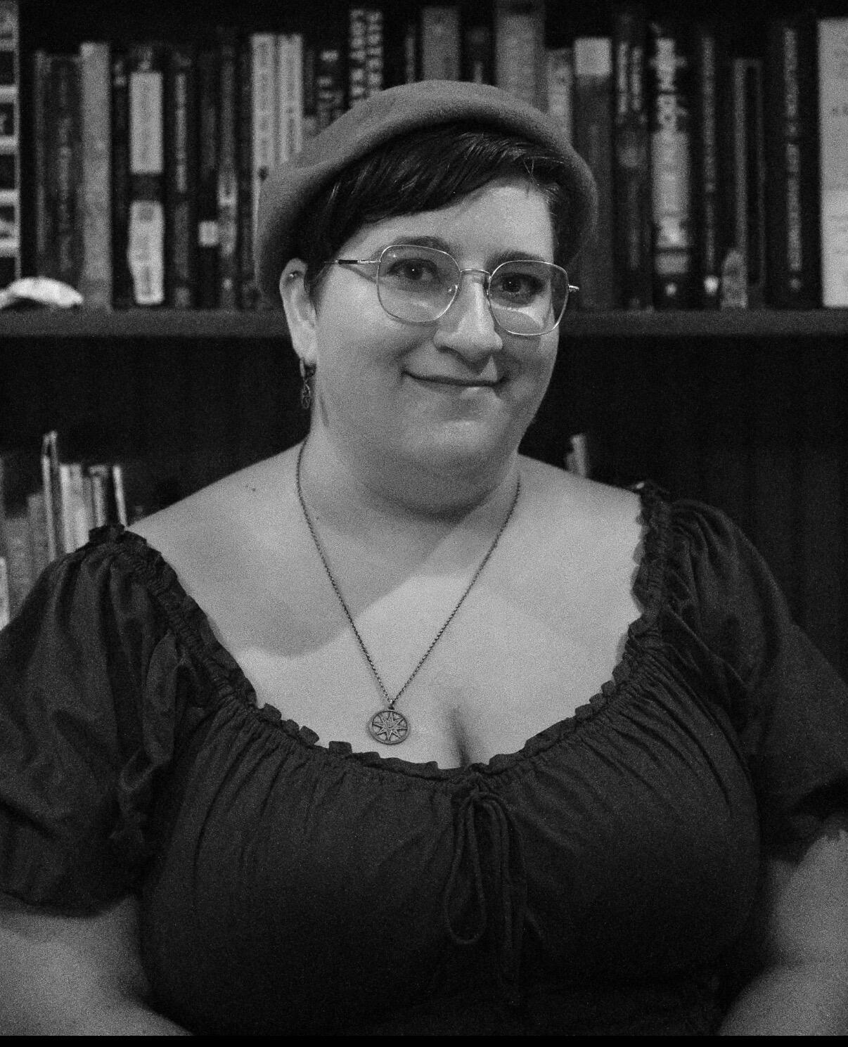 Head-and-shoulders picture of a white person with short, dark brown hair, wearing octagonal glasses, a stripped shirt, and a silver necklace, against a light gray background fading to black. They are smiling at the camera.